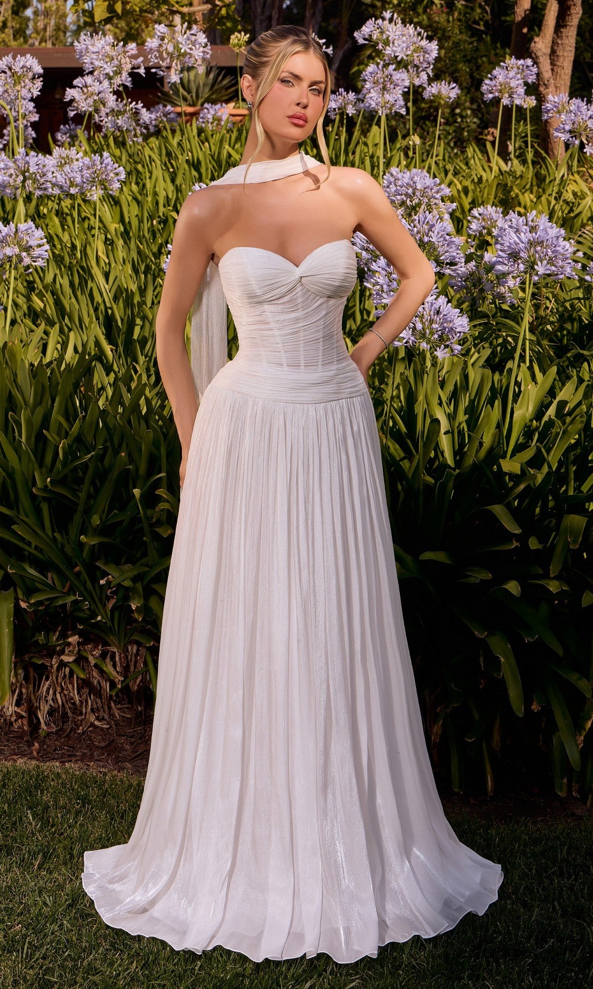 Standing outdoors in a garden surrounded by purple flowers and greenery, the woman wears the Ladivine CD414W long formal dress in off white, featuring a strapless fitted corset bodice and pleated skirt, complemented by a white neck scarf.
